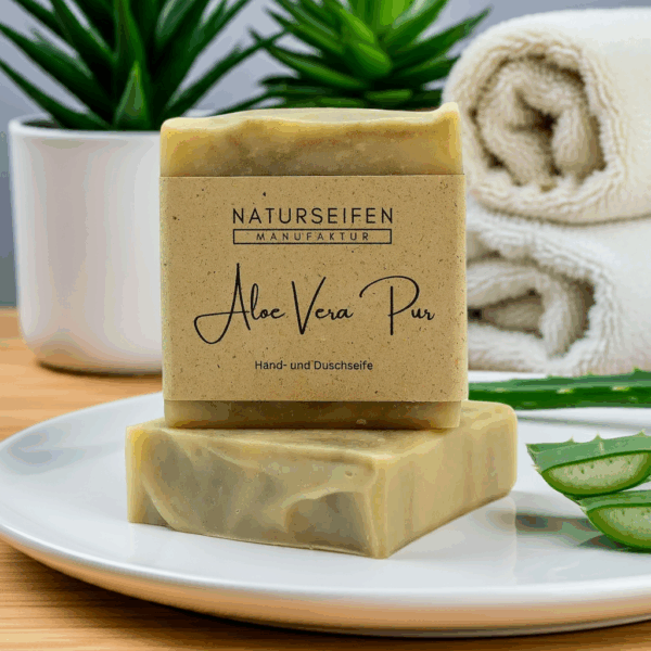 Zwei Stücke Aloe Vera Seife mit einer Verpackung auf einem Teller neben Handtüchern und Pflanzen.