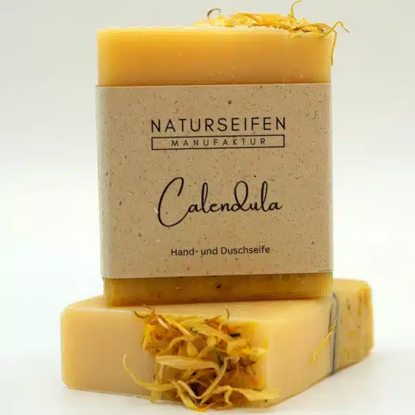 Two pieces of handmade calendula soap from Naturseifen Manufaktur, one stacked on top of the other. The soap is yellowish and has calendula flowers incorporated into it. It has a natural-colored banderol with the inscription "Naturseifen Manufaktur Calendula hand and shower soap".