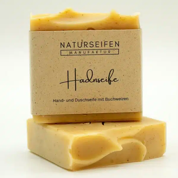 Zwei Stücke handgefertigte Hadnseife von Naturseifen Manufaktur, eine übereinander gestapelt. Die Seife ist beige mit kleinen braunen Punkten und hat eine naturfarbene Banderole mit der Aufschrift "Naturseifen Manufaktur Hadnseife Hand- und Duschseife mit Buchweizen".