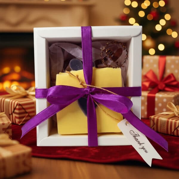 Eine weiße Geschenkbox für 1 Naturseife mit lilafarbener Schleife und Dankeschön-Anhänger steht inmitten verpackter Geschenke, während im Hintergrund ein Kamin und ein geschmückter Weihnachtsbaum warm leuchten.