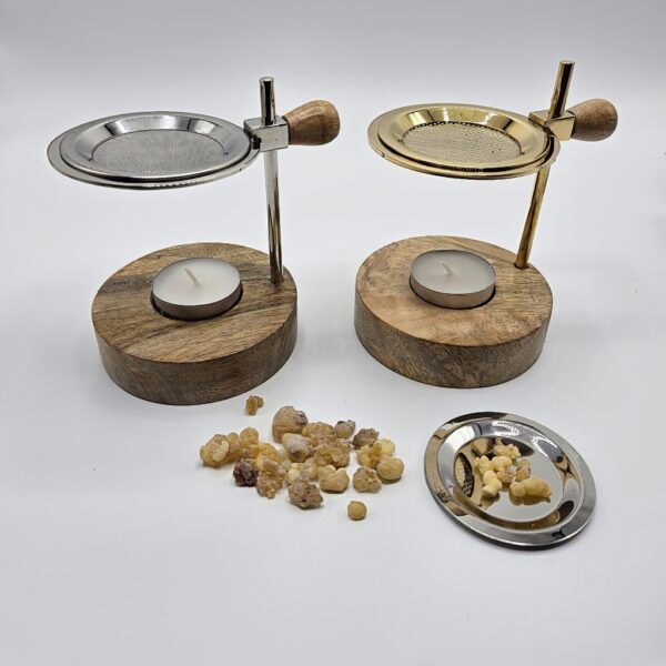 Two incense burners with wooden bases and tea lights. One has a silver sieve, the other a gold one. In front of them are pieces of incense and a small plate with resin.