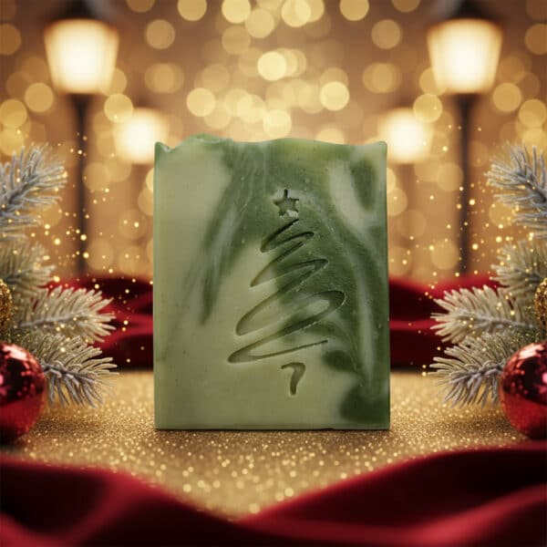 Die Aloe Vera Naturseife "X-Mas Edition" ist ein festliches grünes Seifenstück mit Weihnachtsbaummotiv auf rotem Samt, umgeben von Tannenzweigen, rotem Schmuck und leuchtenden Lichtern, die im Hintergrund verschwimmen.