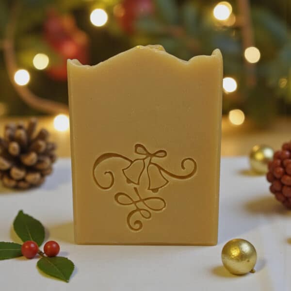 Ein handgefertigtes Seifenstück mit einem dekorativen Glockenmuster steht aufrecht auf einer weißen Fläche. Tannenzapfen, Stechpalmenblätter, Beeren und goldene Ornamente umgeben es. Warme Lichterketten und festliches Grün verschwimmen im Hintergrund.
